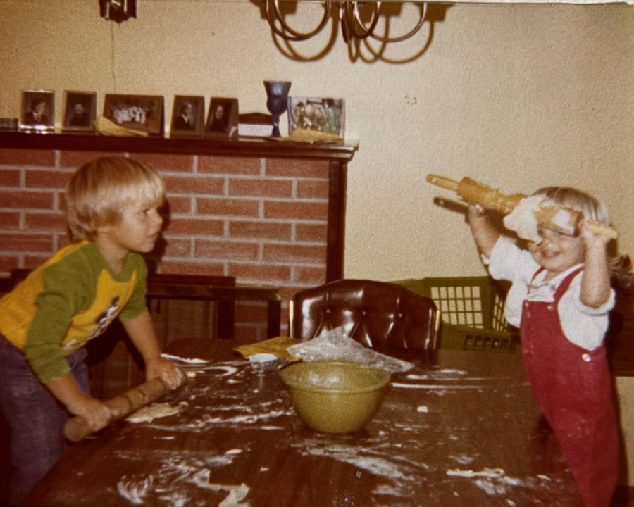 Photo is from the 1970s. It shows a young boy and girl standing on chairs alongside a table. The table is covered in flour, and the children are playing with rolling pins and dough.