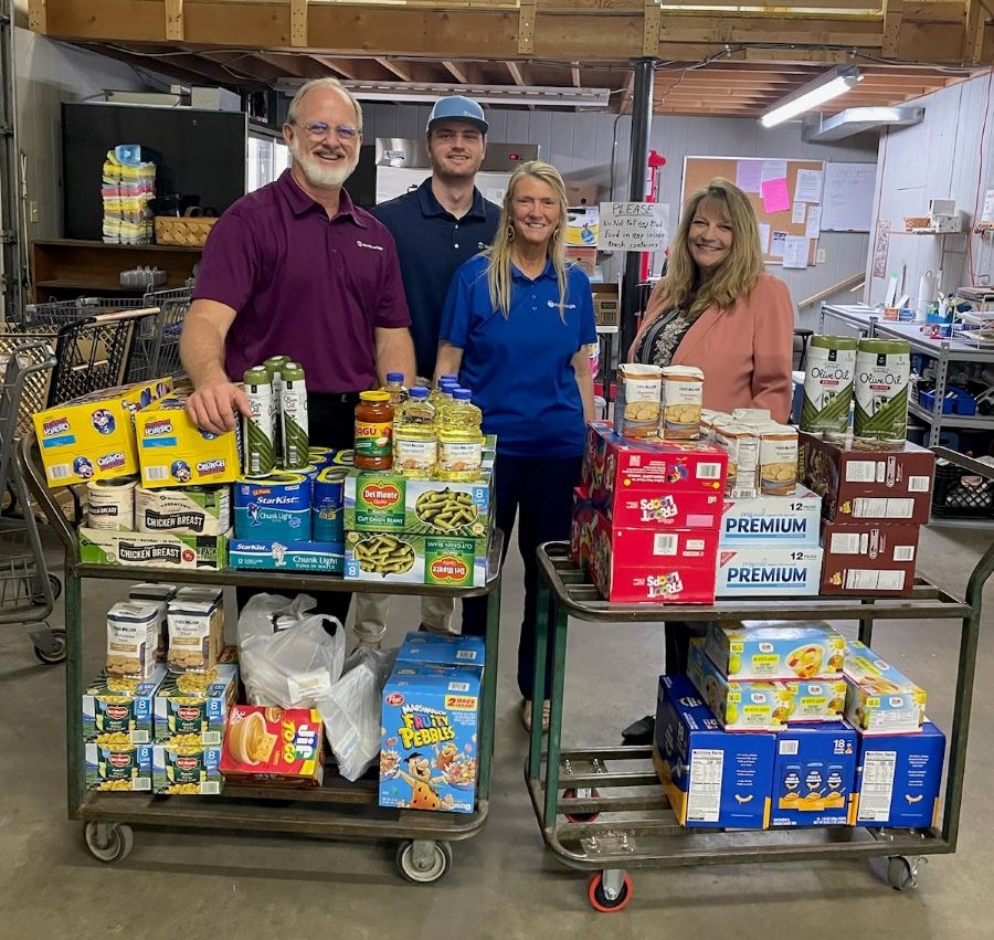 Four people stand behind stand behind wheeled carts filled with food. Among the food is canned chicken, tuna, green beans, corn, cereal, boxed mac and cheese, crackers, and olive oil.