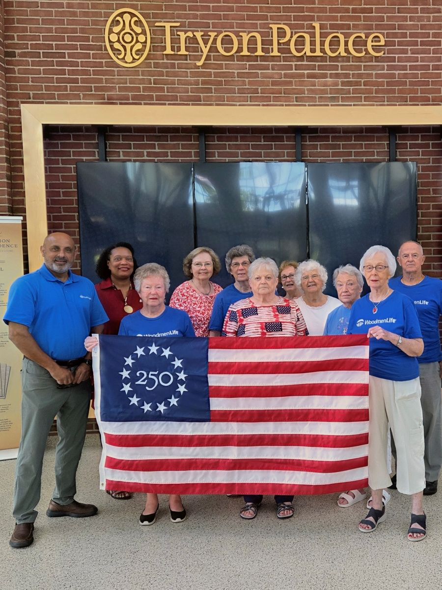 Photo shows 11 people standing together holding a commemorative Betsy Ross America 250 flag. Behind them is a brick building with the words "Tryon Palace" above them.