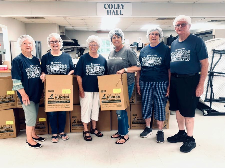 Six people appear in the photo. They are all wearing WoodmenLife-branded T-shirts and hairnets. There are cardboard boxes in their hands and piled behind them that say "Feed the Hunger, Beans and Rice Meal Packets, Humanitarian Relief."