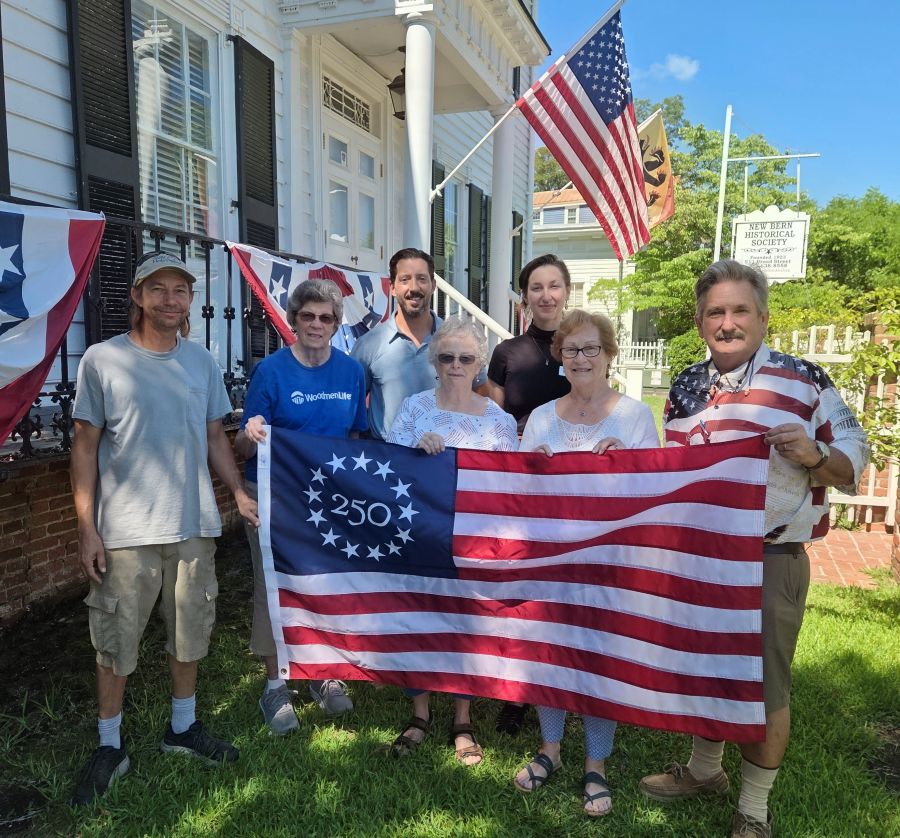Photo shows seven people standing together holding a commemorative Betsy Ross America 250 flag. They are standing outside, and behind them is a white building. There is also a sign behind them that says "New Bern Historical Society."