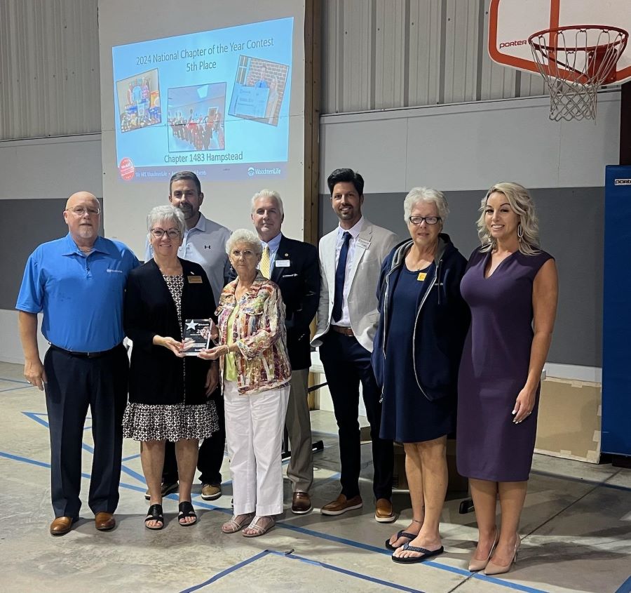 Eight people are standing together. Two of the people in front are holding a glass award. Projected on the wall behind the group is a screen that says "2024 National Chapter of the Year Contest, 5th Place, Chapter 1483 Hampstead."