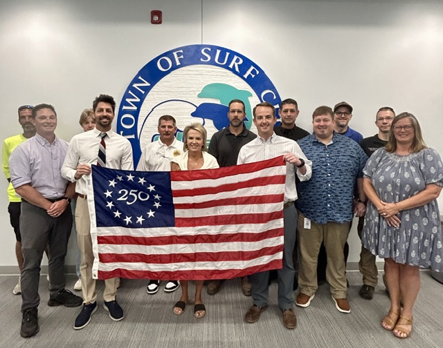Photo shows 13 people standing together holding a commemorative Betsy Ross America 250 flag. Behind them is a white wall with a Town of Surf City logo.
