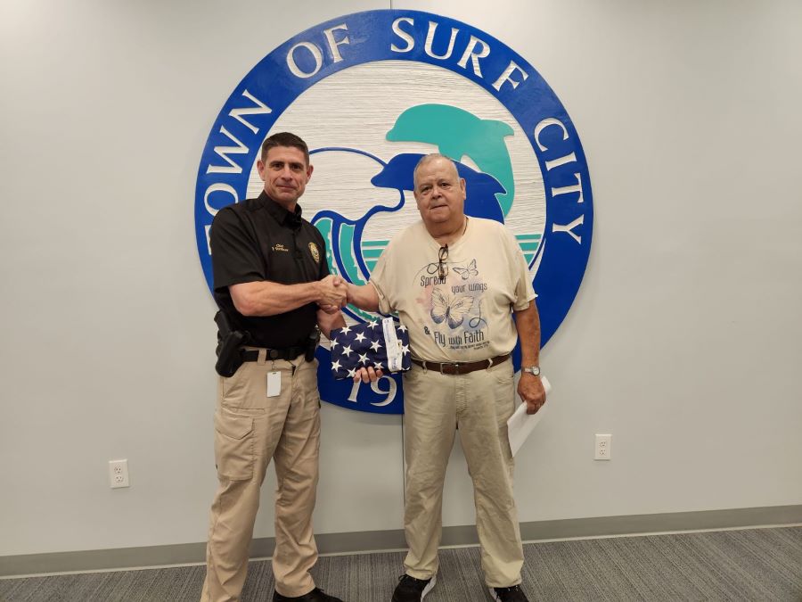 Two men stand shaking hands and holding an American flag. Behind them on the wall is the seal of the town of Surf City, NC.