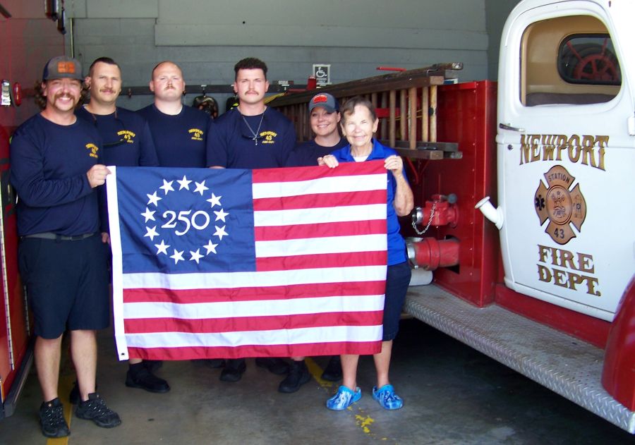 Photo shows six people standing together holding a commemorative Betsy Ross America 250 flag. Five of the people are firefighters and are wearing matching navy blue shirts. To their right is a firetruck that says "Newport Fire Dept., Station 43" on the door."