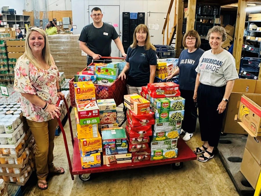 Five people appear in the picture. They are with shopping carts filled with boxed and canned food. They are standing in a warehouse with crates of nonperishable food items.