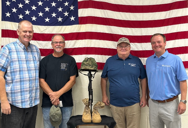 Four men stand together for the photo. Behind them hanging on the wall is an American flag. In the middle of the men is a military helmet and boots.