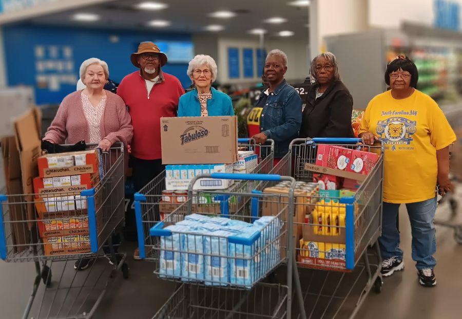 Six people stand behind four grocery carts. The carts are filled with various foods for donation, including ramen packages, canned food, and condiments.