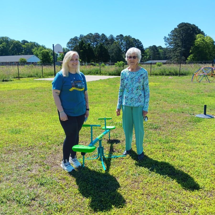 Two women stand alongside a blue-and-green teeter totter. they are standing outside; behind them is lots of green grass and a small basketball court.