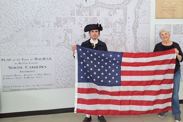 A man and a woman pose holding an American flag. The man is wearing historic clothing. On the wall behind them is a large black-and-white map of Halifax, NC.