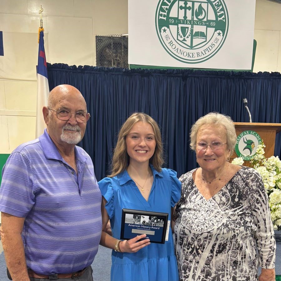 An older man and woman stand on either side of a student. The student is holding a plaque that says "American History Award." Behind them is a stage with a podium and "Halifax Academy, Vikings" branding.