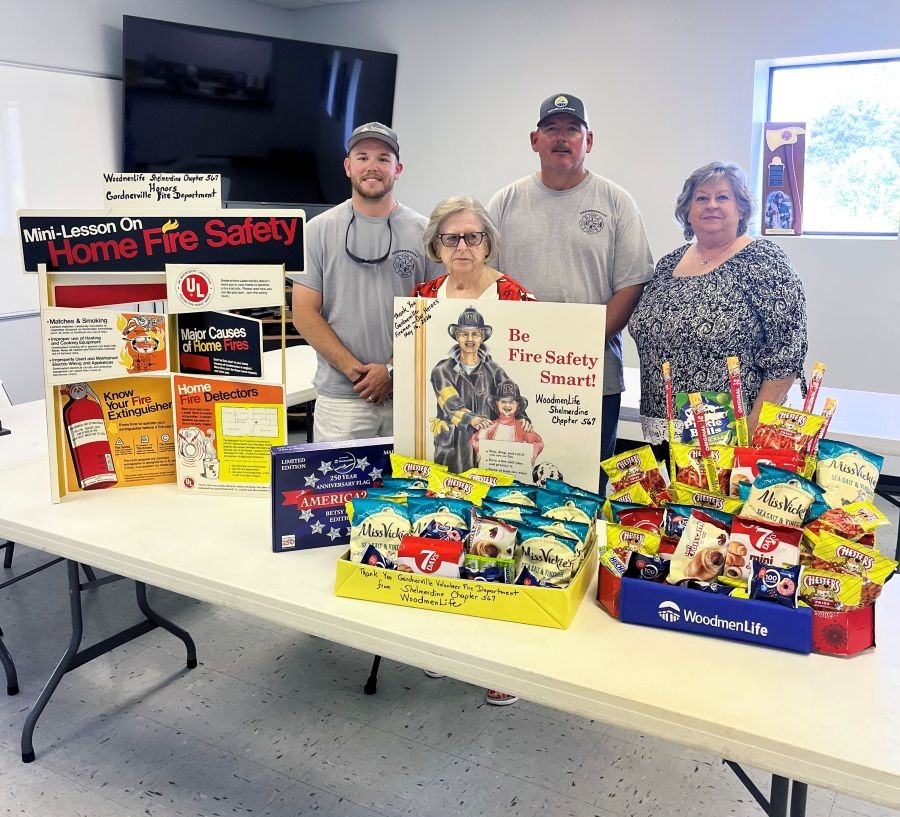 Four people -- two women and two men -- stand together for the photo. They are standing behind a table. On the table are boxes full of chips and other snacks. Also on the table are display posters about fire safety.