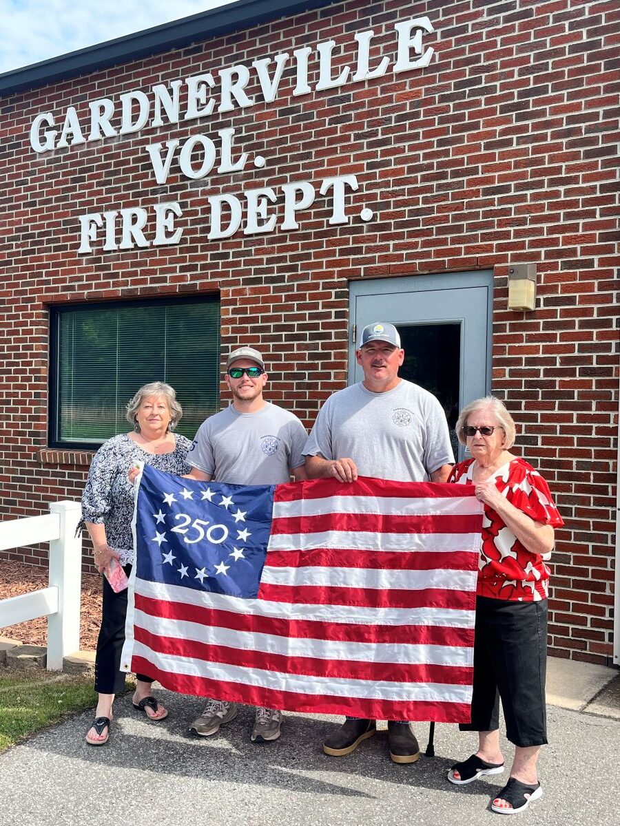 Four people -- two women and two men -- stand together for the photo. They are holding a Betsy Ross 250 commemorative flag. On the flag, the blue corner says "250" and has a circle of stars. The group is standing outside a brick building that says "Gardnerville Vol. Fire Dept."