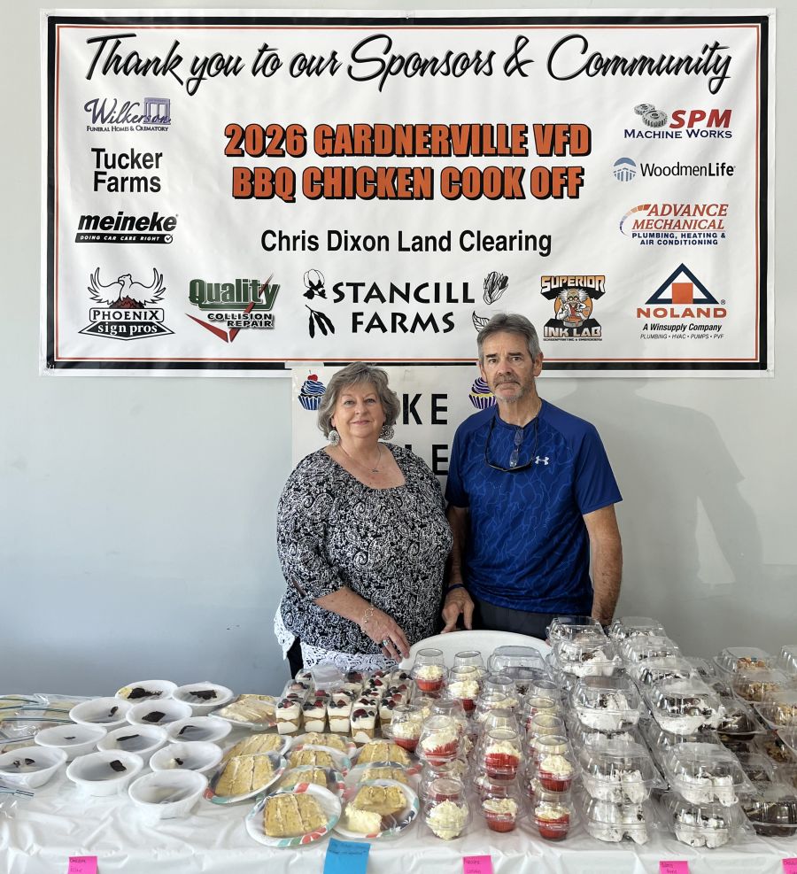 A man and a woman stand together for the photo. In front of the people is a table with baked goods: slices of cake on plates, packages of cupcakes, etc. Behind them is a sign hanging on the wall that says "Thank you to our Sponsors & Community 2026 Gardnerville VFD BBQ Chicken Cook Off," with several business logos.