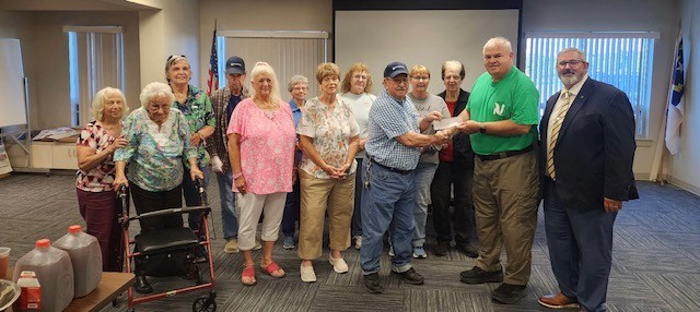 13 people stand together for the photo. Two men near the right side are exchanging a check and shaking hands.