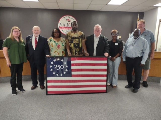 Eight people stand together for the photo. They are holding a framed America 250 commemorative flag, which features 13 stars in a circle around "250" in the upper-left blue square. Behind them is a long desk, and on the wall is the seal of the town of Warsaw, NC.