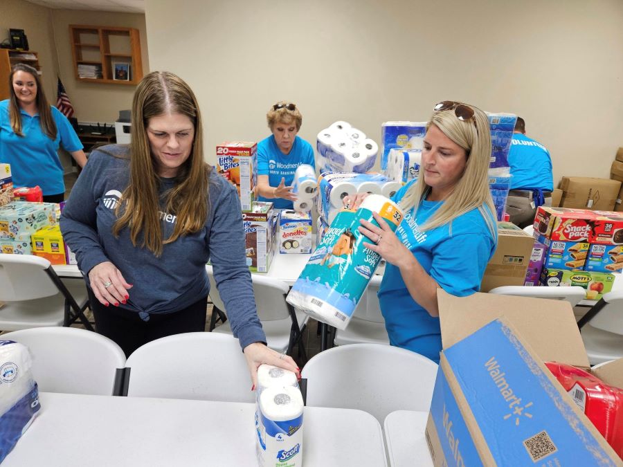 Four women are organizing donations on tables. Among the donations are toilet paper and shelf-stable food.