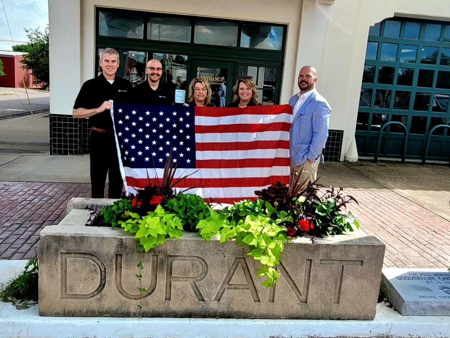 A group of five people stand holding an American flag. In front of them is a large cement planter that says "Durant" on the front. The group is standing outside.