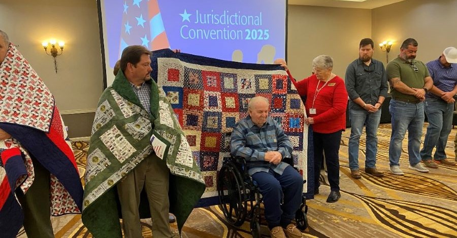 A man in a wheelchair is at the center of the photo. People are holding a quilt up behind him, and it is about to be draped over his shoulders. Men are lined up on either side of him -- the men on the left already have quilts, and the men on the right are about to receive their quilts.