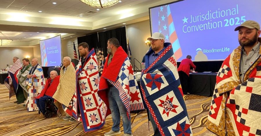 Photo shows nine men standing in a row. Each of the men has a quilt wrapped around their shoulders. Behind them is a large projector screen that says "Jurisdictional Convention 2025," with the WoodmenLife logo at the bottom.