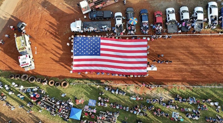Photo is a drone shot looking down on an oversized American flag. People are standing around the edge of the flag holding it. They are holding it open on a wide dirt path. Alongside the dirt path are vehicles and spectators watching.
