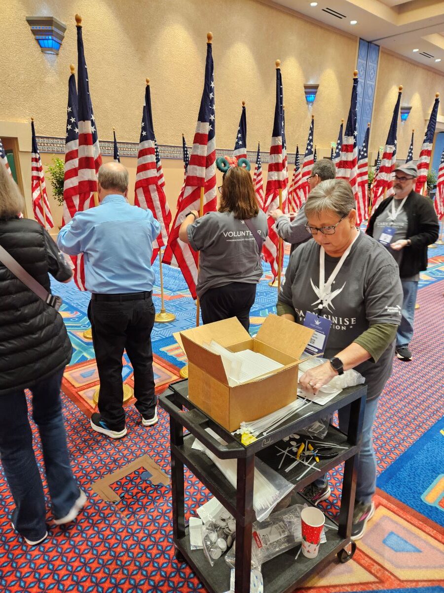 Several people stand among rows of flags standing upright on flagpoles. The volunteers, who are wearing dark gray Gary Sinise Foundation T-shirts, and straightening the flags.