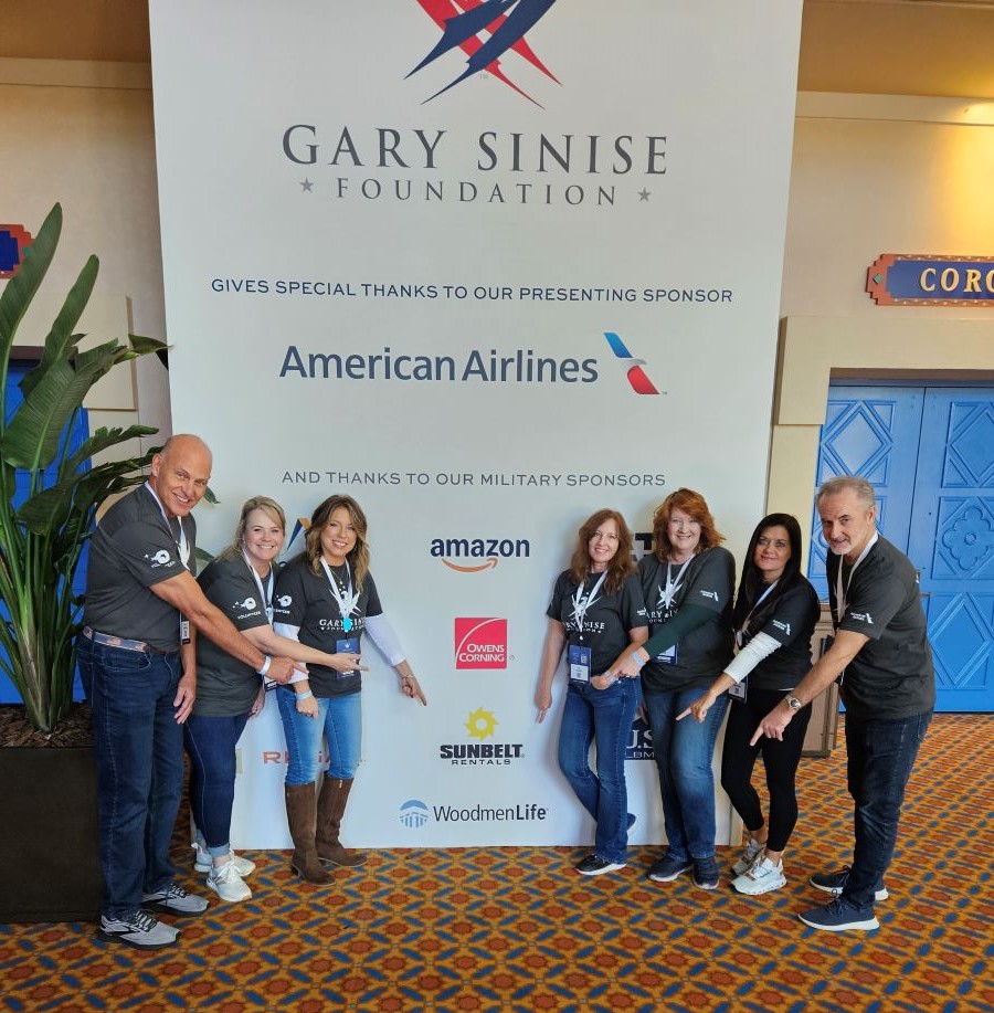 Seven people are in this photo. They are flanking a large sign that says "Gary Sinise Foundation gives special thanks to our presenting sponsor American Airlines and thanks to our military sponsors." Below those words are several logos of companies sponsoring the Snowball Express Annual Event. The people in the photo are all pointing to the WoodmenLife logo, which appears at the bottom.