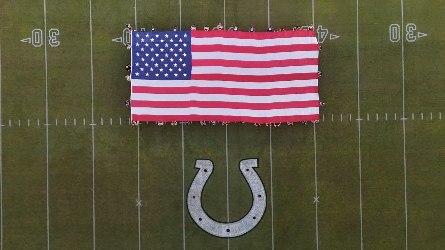 Photo shows a drone image looking down on an oversized American flag displayed on a football field. There is a horseshoe painted on the field below the flag.
