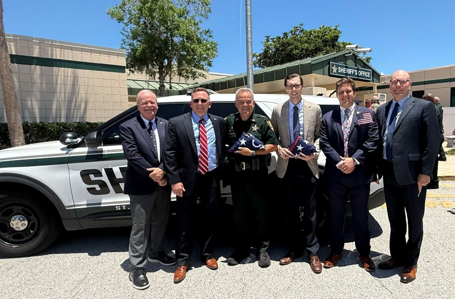 Six men stand together outside a Sheriff's Office. They are holding two folded-up commemorative Betsy Ross 250 U.S. flags.