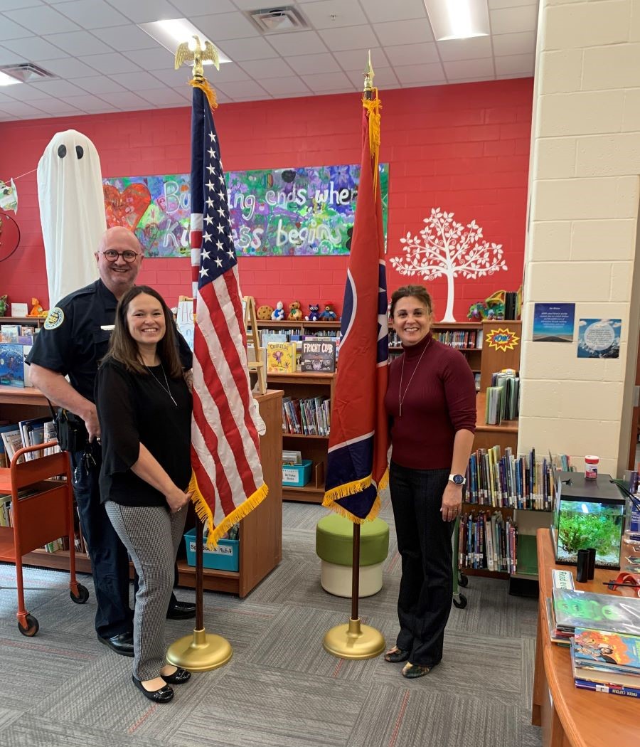 Three people stand in a school library alongside a U.S. flag and a Tennessee state flag.