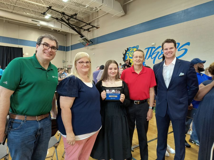 A group of five people stand together in a school gym. At the center is eighth-grade student Lydia Wages; she holds a plaque noting her American History Award. She is flanked by Leah and Mike Wages. On the far left and right sides of the group are WoodmenLife Community Outreach Advisor Scott Murray and Regional Director Brad Lowe.