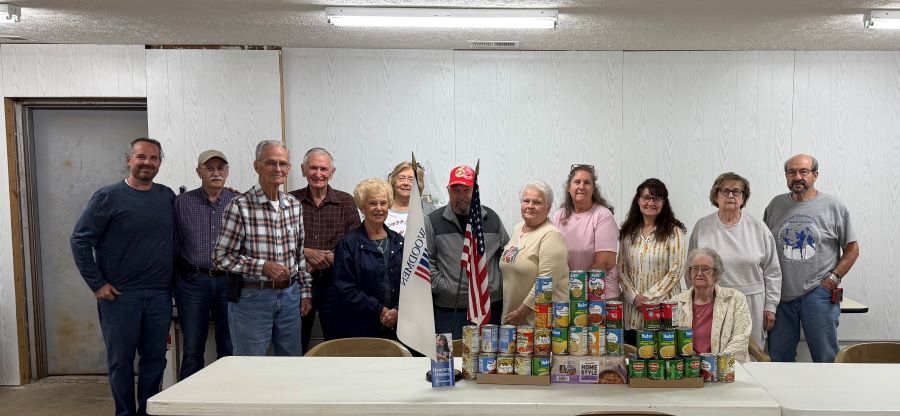 13 people stand behind a long white table. On the table are stacks of canned food, plus an American and a Woodmen of the World flag.