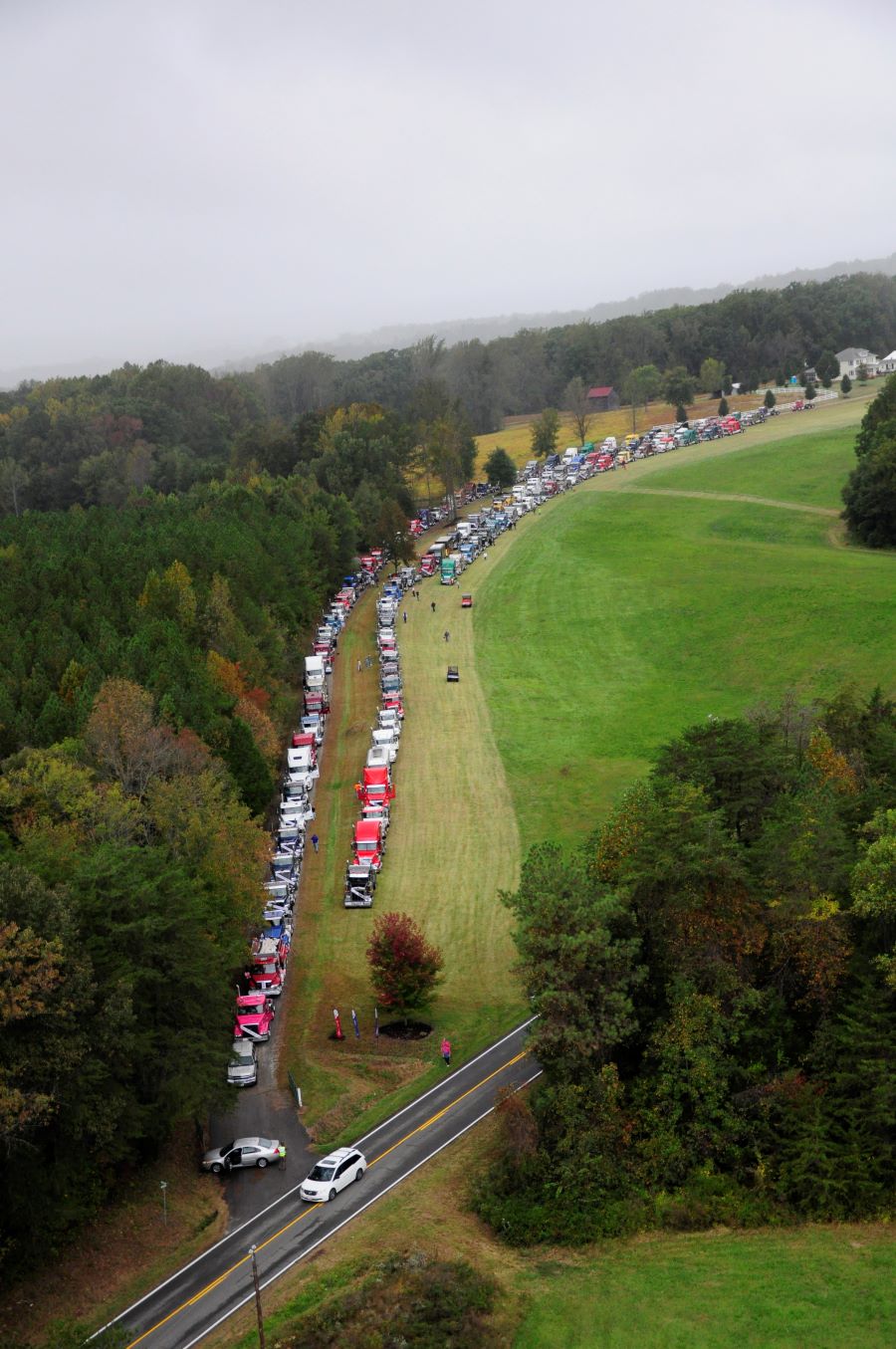 The photo is shot from high overhead. It shows long rows of semitrucks lining up for the Truckers Parade Against Cancer (TPAC). The surrounding area is very green with lots of trees.