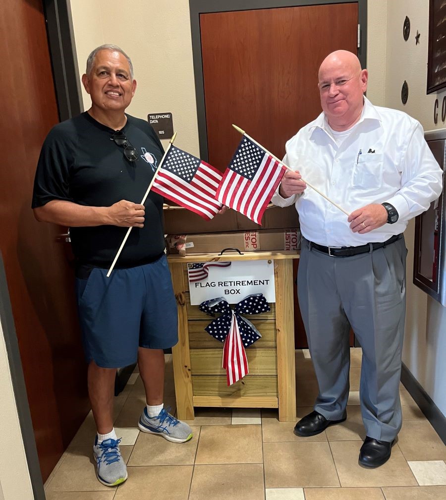 Two men stand together, each holding a handheld flag. They are standing next to a box that says "Flag Retirement Box."