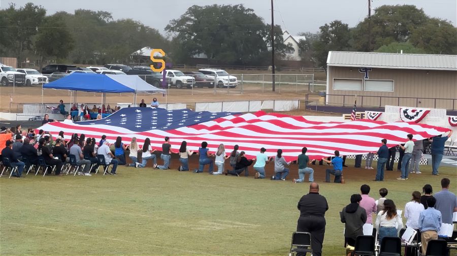 Image shows an oversized American flag on display on an outdoors athletic field. People are surrounding the flag and holding it up — the people in the back are standing, and the people in the front are kneeling, so the flag is slightly tilted for a better view in the audience. And audience of people in folding chairs are in front of the flag.