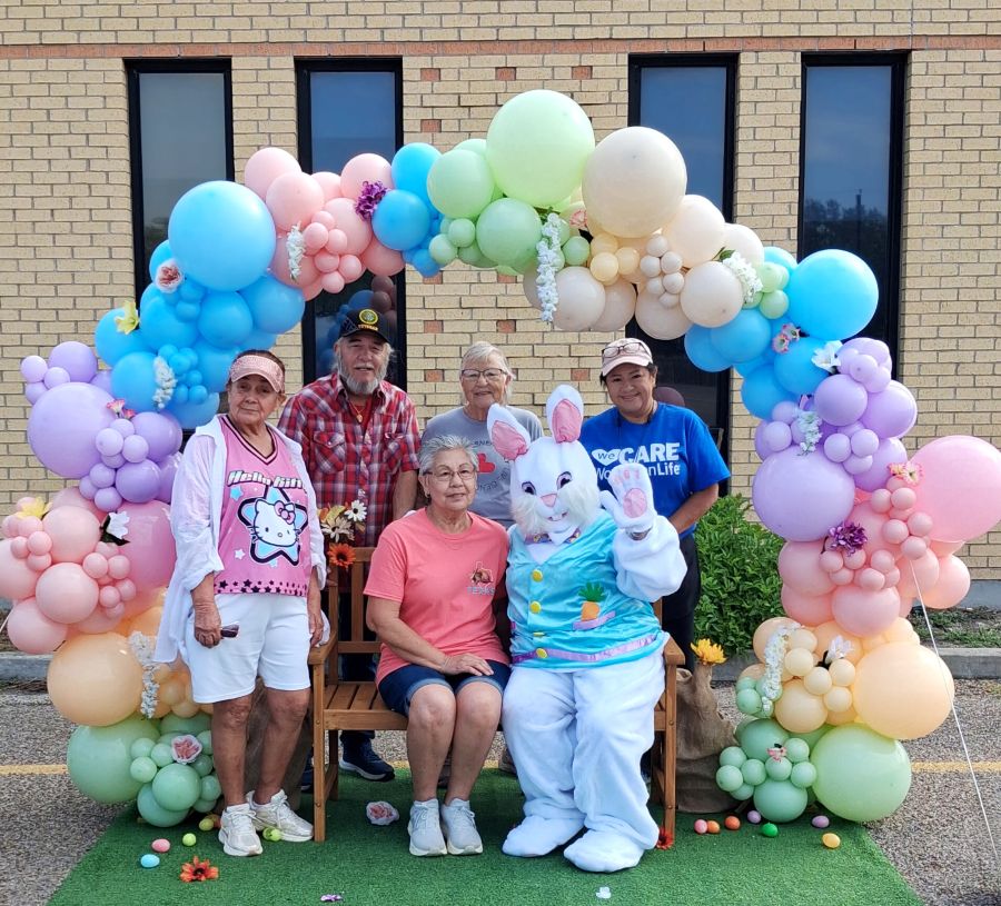 Five people pose for a picture with someone in an Easter bunny costume. They are standing/sitting below a balloon arch, made up of pastel-colored balloons.