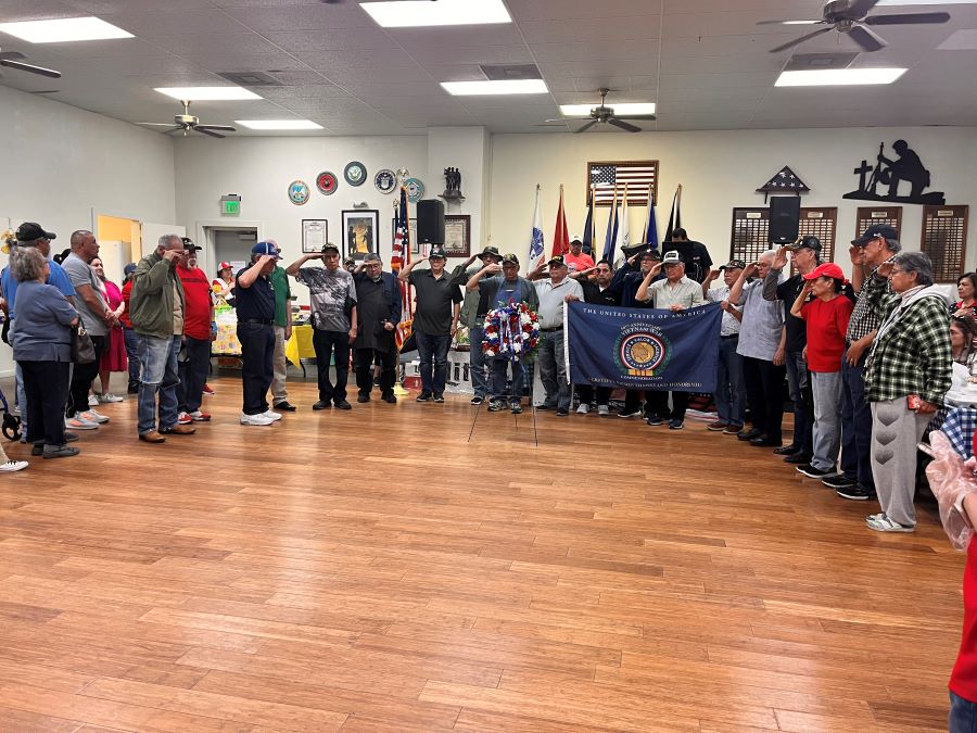 A group of people stand around an American Legion hall. Most people are saluting. At the center of the group is a Vietnam War flag and a red, white and blue wreath.