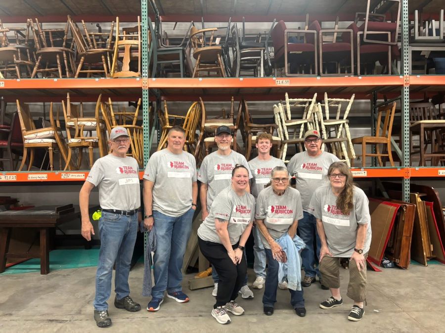 A group of eight people stand together for the photo. They are all wearing grey "Team Rubicon" T-shirts. Behind them are several shelves filled with chairs.