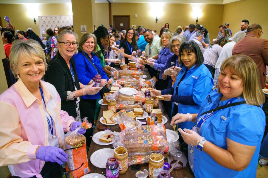 Photo is looking down a long table. People are gathered on either side of the table making peanut butter and jelly sandwiches.