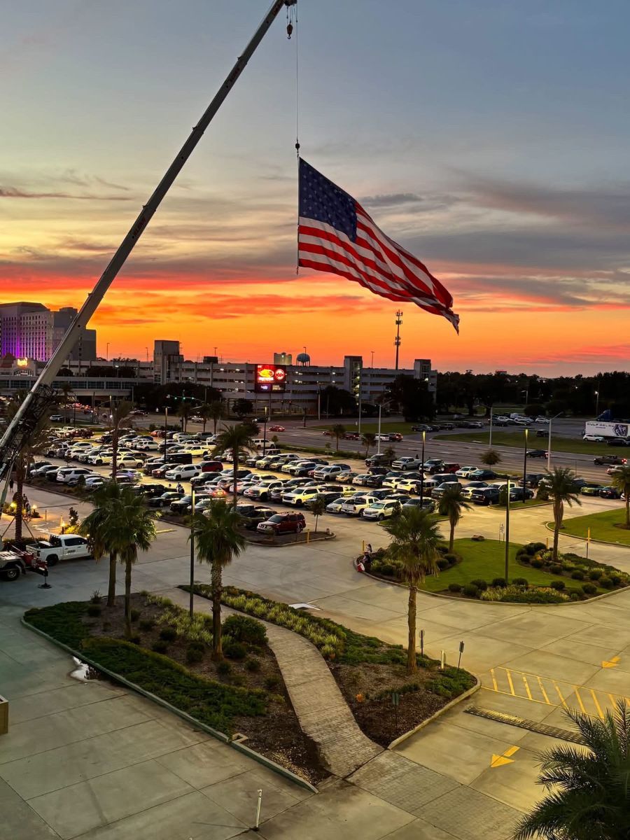 Photo shows an oversized American flag hoisted up by a crane. In the background is a parking lot with the setting sun behind it.