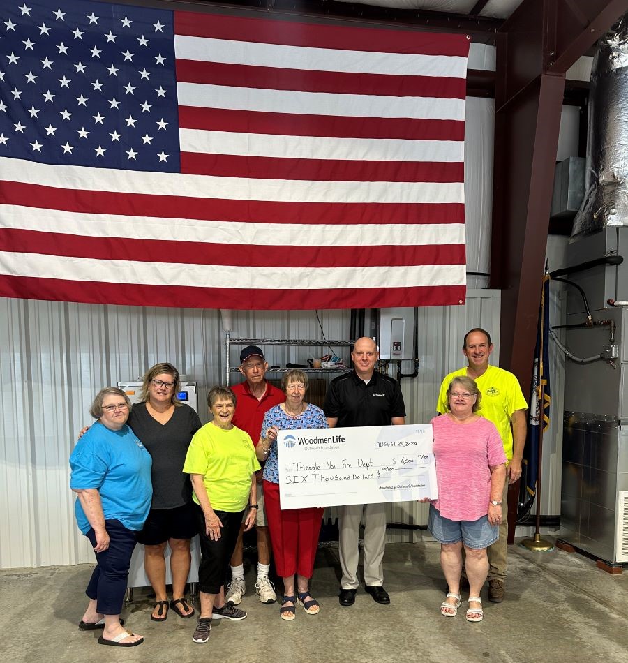 A group of eight people stand together holding an oversized check. The group stands below a large American flag.