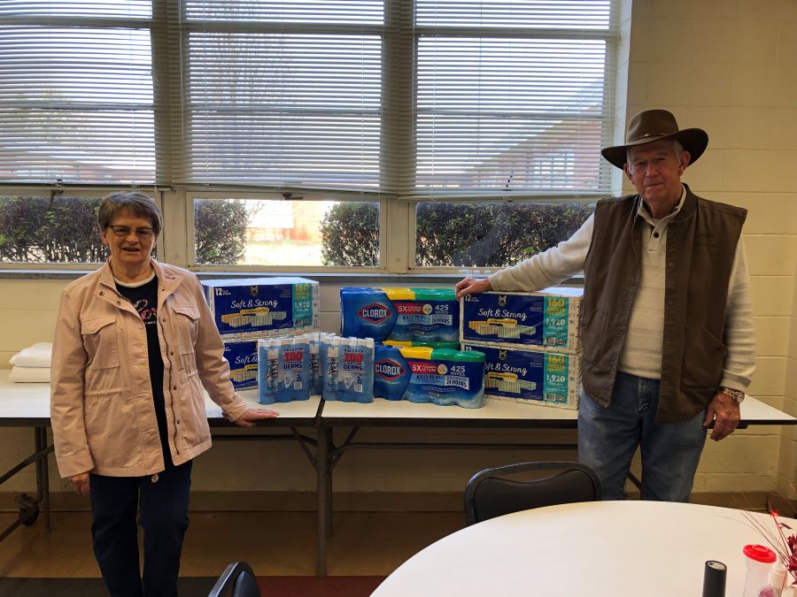 A man and a woman stand alongside a table. Stacked on the table are the donated cleaning supplies, including Lysol spray, Clorox wipes, and boxes of Kleenex.