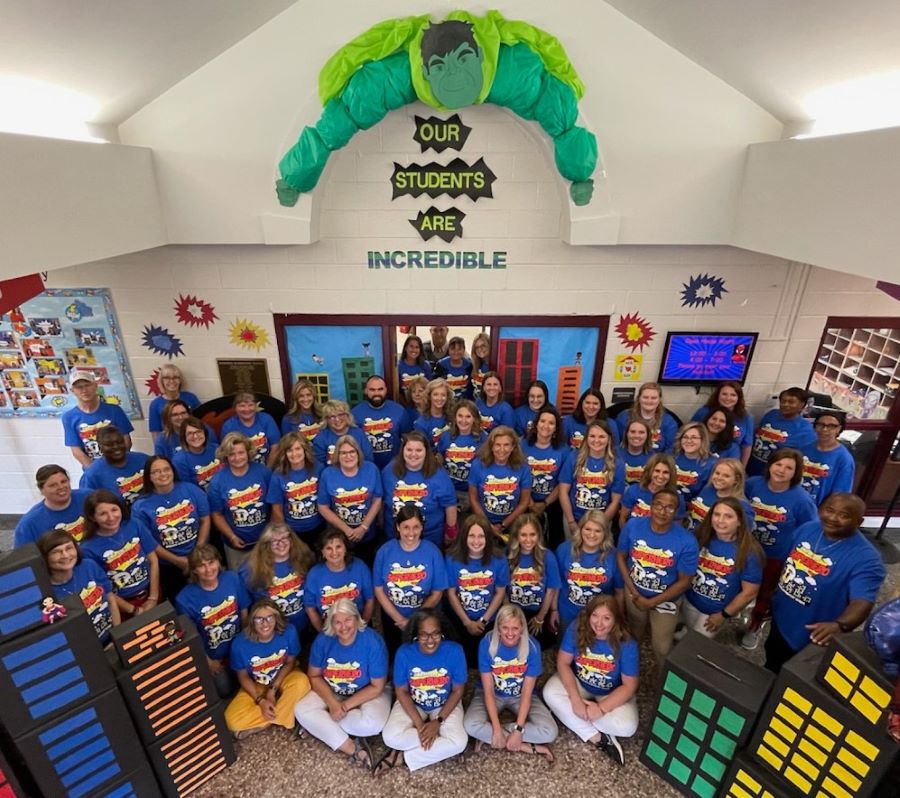 A group of more than 50 teachers, staff and faculty pose for a photo. They are all wearing matching blue T-shirts. On the wall above them is an image of the Incredible Hulk, with the words "Our Students Are Incredible."