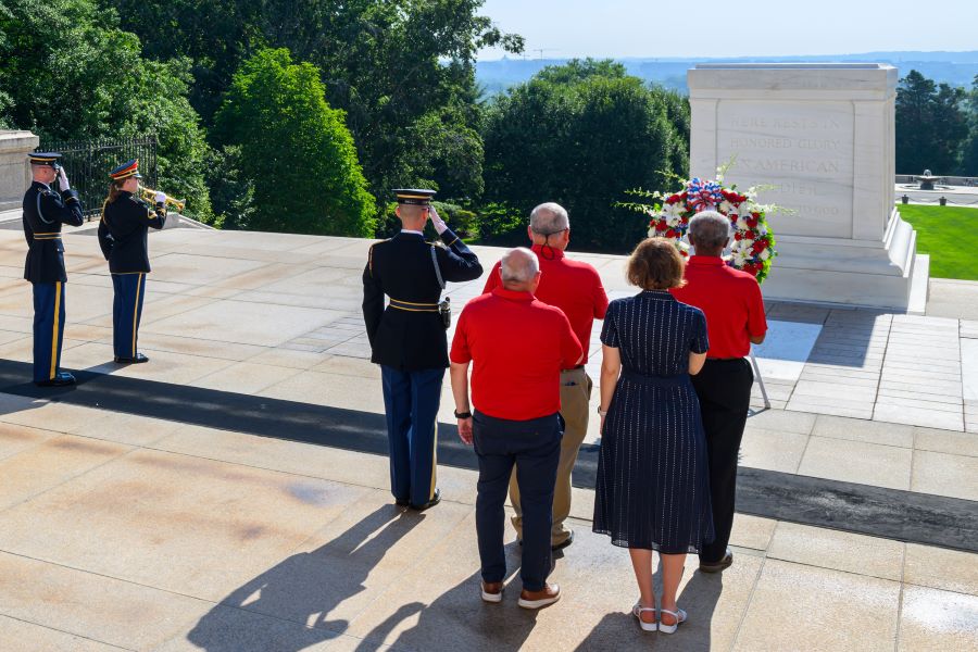 A group of four people stand with their backs to the camera. They are facing the Tomb of the Unknown Soldier at Arlington National Cemetery. In front of them is a wreath propped up on a stand. Three service members are standing at attention.