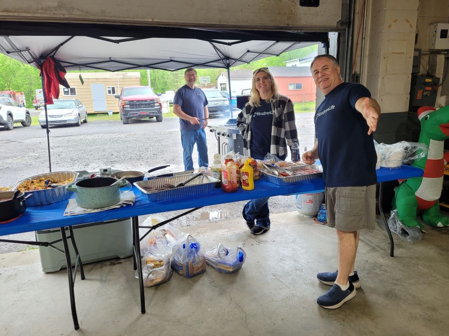 Three people, all wearing navy blue WoodmenLife T-shirts, stand at folding tables. On the tables are pans filled with hamburgers, hot dogs and other food.