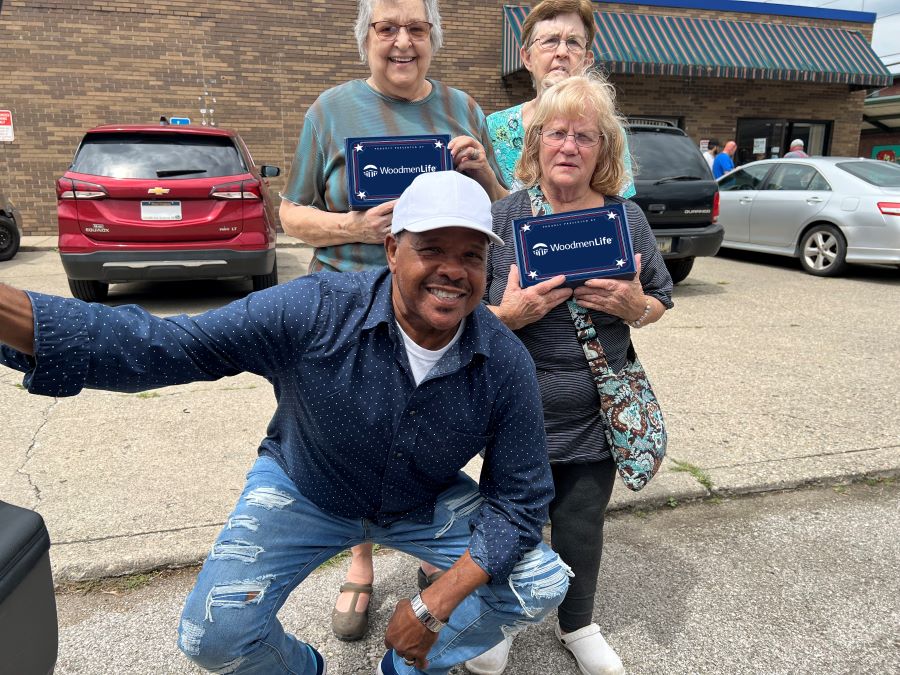 Four people are in the photo. The three women — all WoodmenLife members — are presenting flag boxes to the man. The group is standing outside.