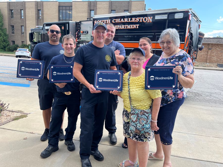 Seven people — a mixture of WoodmenLife members and firefighters — stand together holding four WoodmenLife-branded flag boxes. The group is outside, and there is a firetruck parked behind them.