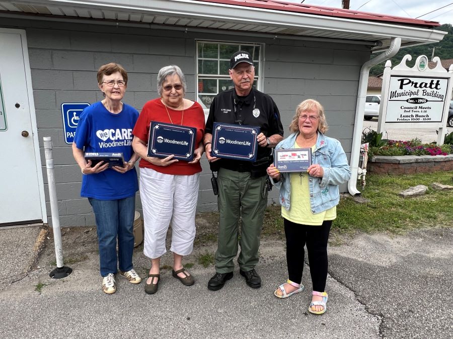 Three women stand with a male police officer. Each of the four people is holding a WoodmenLife-branded flag box. They are standing outside the Pratt municipal building.