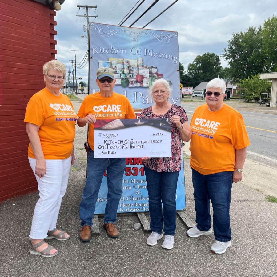 Photo shows four people standing together holding an oversized check. The WoodmenLife-branded check is made out to Kitchen of Blessing in the amount of $1,500. Three of the people are wearing orange shirts that say "WeCare, WoodmenLife, Fight Hunger." They are standing outside in front of a sign that says "Kitchen of Blessing."