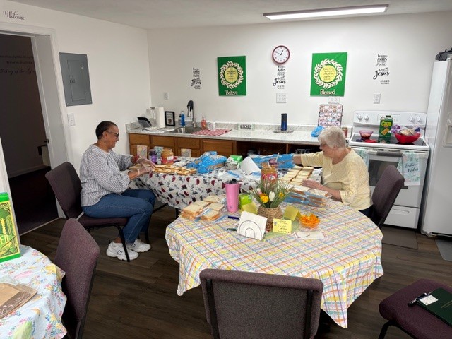 Photo shows two women seated at a table. Spread out on the table in front of them are slices of bread and open containers of peanut butter and jelly.